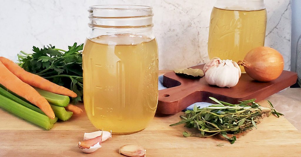 This is an image of two containers of vegetable broth staged with ingredients, including onions, garlic, carrots, celery, and herbs.