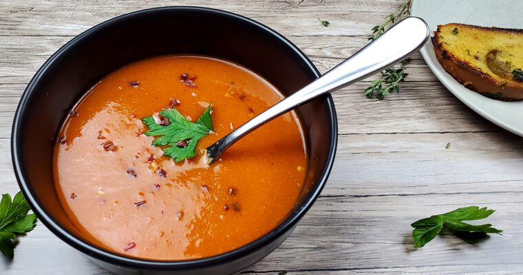 This is an image of a bowl of roasted red pepper chickpea soup garnished with red pepper flakes and fresh parsley. Served with garlic confit toast