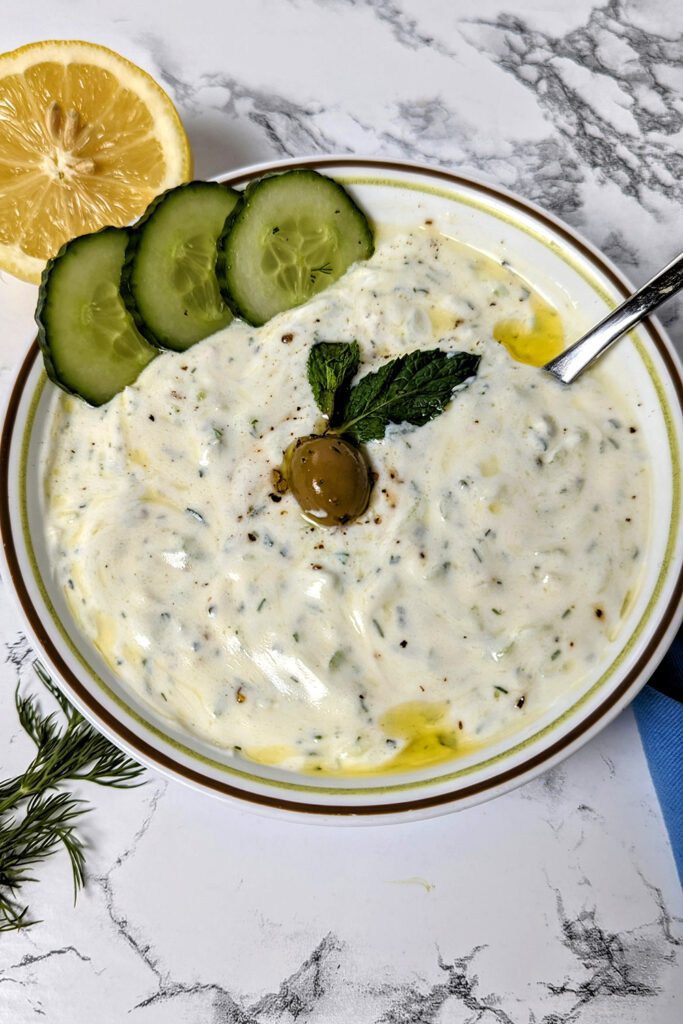 This is an image of the Greek Tzatziki by Sundays at T's. The tzatziki is garnished with cucumbers, fresh mint, and a Kalamata olive.