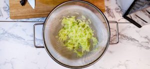 This is an image of grated cucumber with a pinch of salt in a colander sitting over a bowl. Drain the water from the cucumber before adding to your yogurt for a delicious Greek tzatziki.