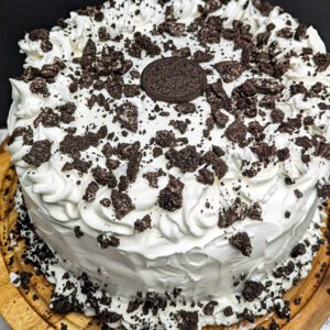 This is an image of the gluten-free 5-layer chocolate ice cream cake. This death by chocolate ice cream cake is made with a gluten-free chocolate cake, gluten-free Oreos, chocolate ice cream, chocolate syrup, Reese's peanut butter ice cream topping, and whipped cream. This image shows a the whole cake frosted with whipped cream and topped with crumbled gluten-free Oreos.