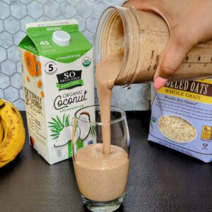 This is an image of the chocolate chia banana smoothie from Sundays at T's. The smoothie is being poured into a class and the image is staged with So Delicious Organic Coconut milk, Bob's Red Mill gluten-free rolled oats, and bananas.