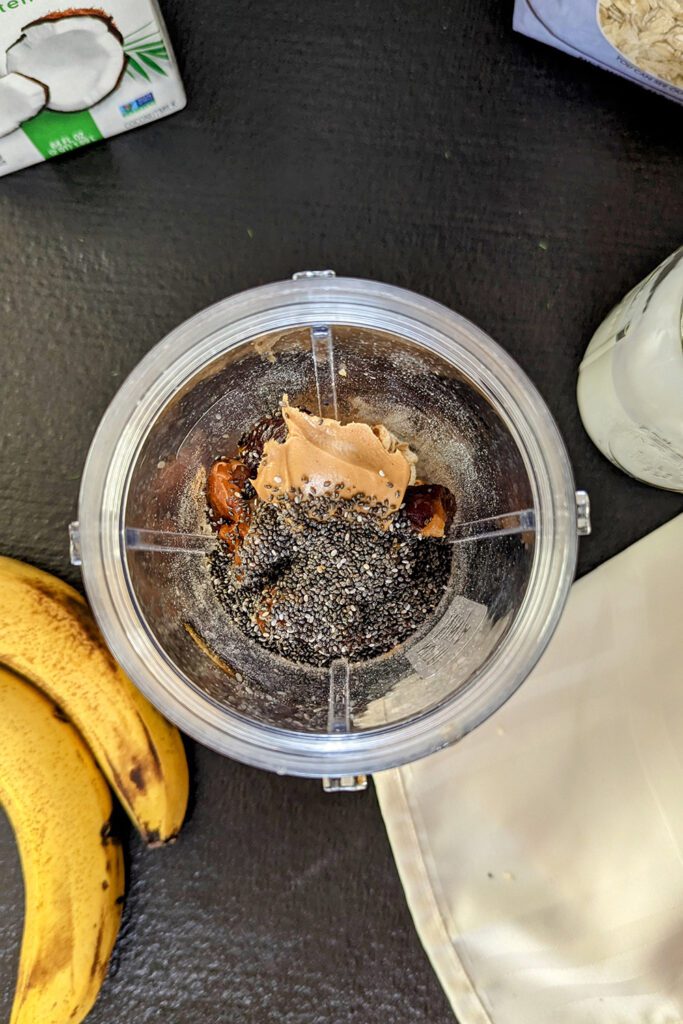 This is an image of the chocolate chia banana smoothie from Sundays at T's. The smoothie ingredients are in the Ninja individual cup ready for blending. The image is staged with So Delicious Organic Coconut milk, Bob's Red Mill gluten-free rolled oats, and bananas.
