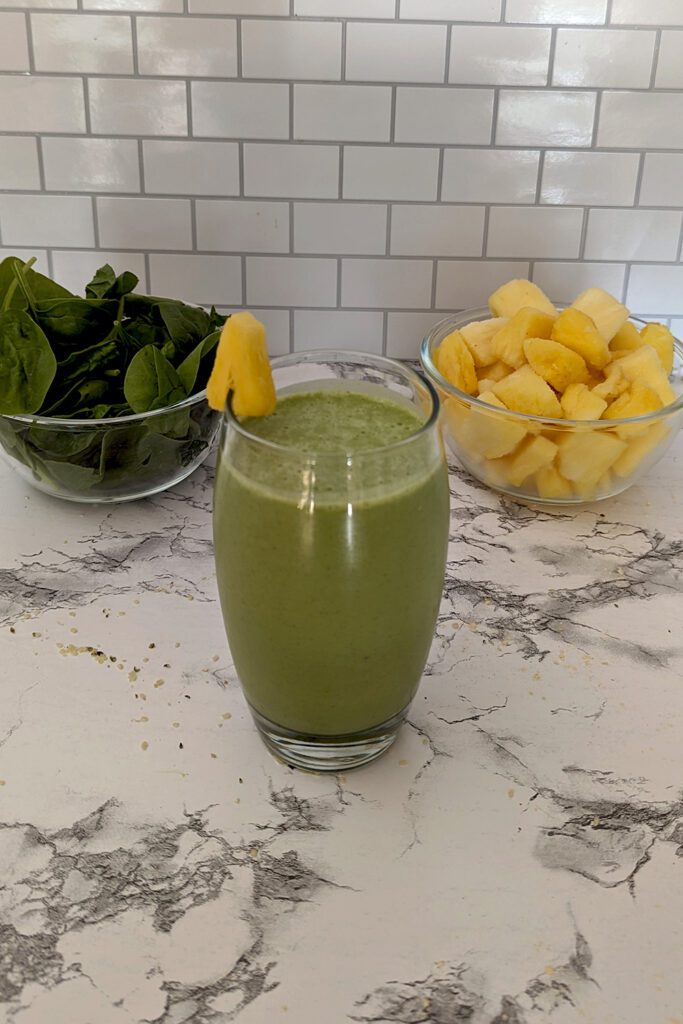 This is an image of the Pineapple Popeye Smoothie from Sundays at T's. The glass is garnished with fresh pineapple. The photo is decorated with spinach and pineapples in the background.